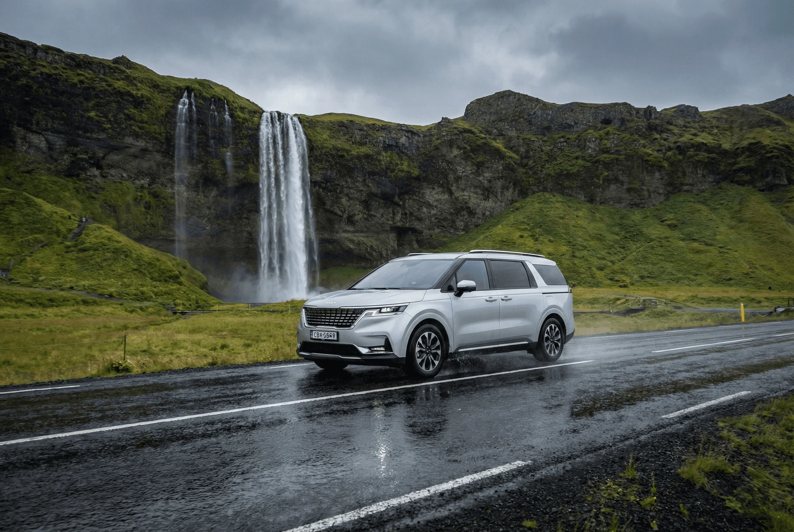 A silver Kia SUV driving on a wet road with a large waterfall and green cliffs in the background.