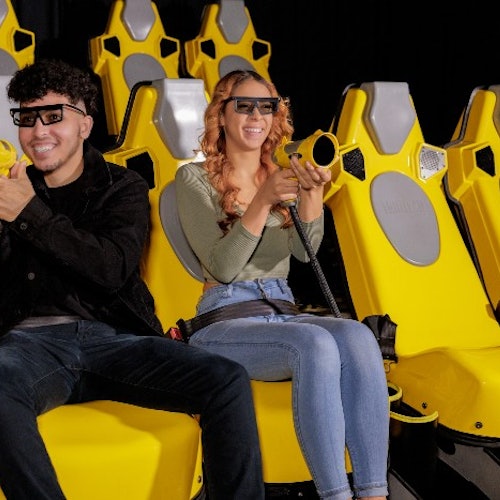 Двама души, носещи 3D очила и държащи лазерни пушки, седят в жълти седалки в тъмна стая, усмихнати и наслаждаващи се на пътешествието.