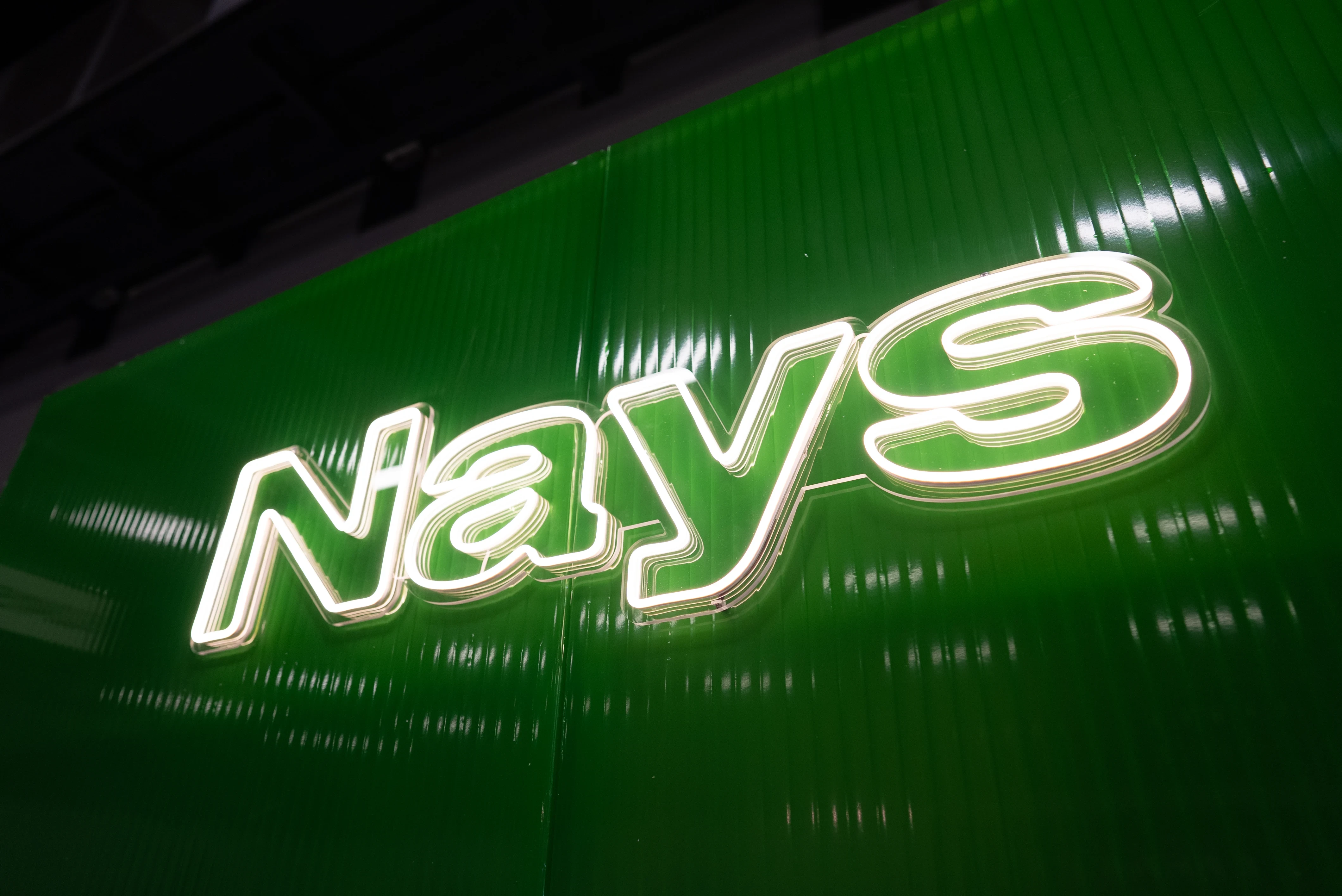 Illuminated Nays neon logo on a glossy green corrugated wall.