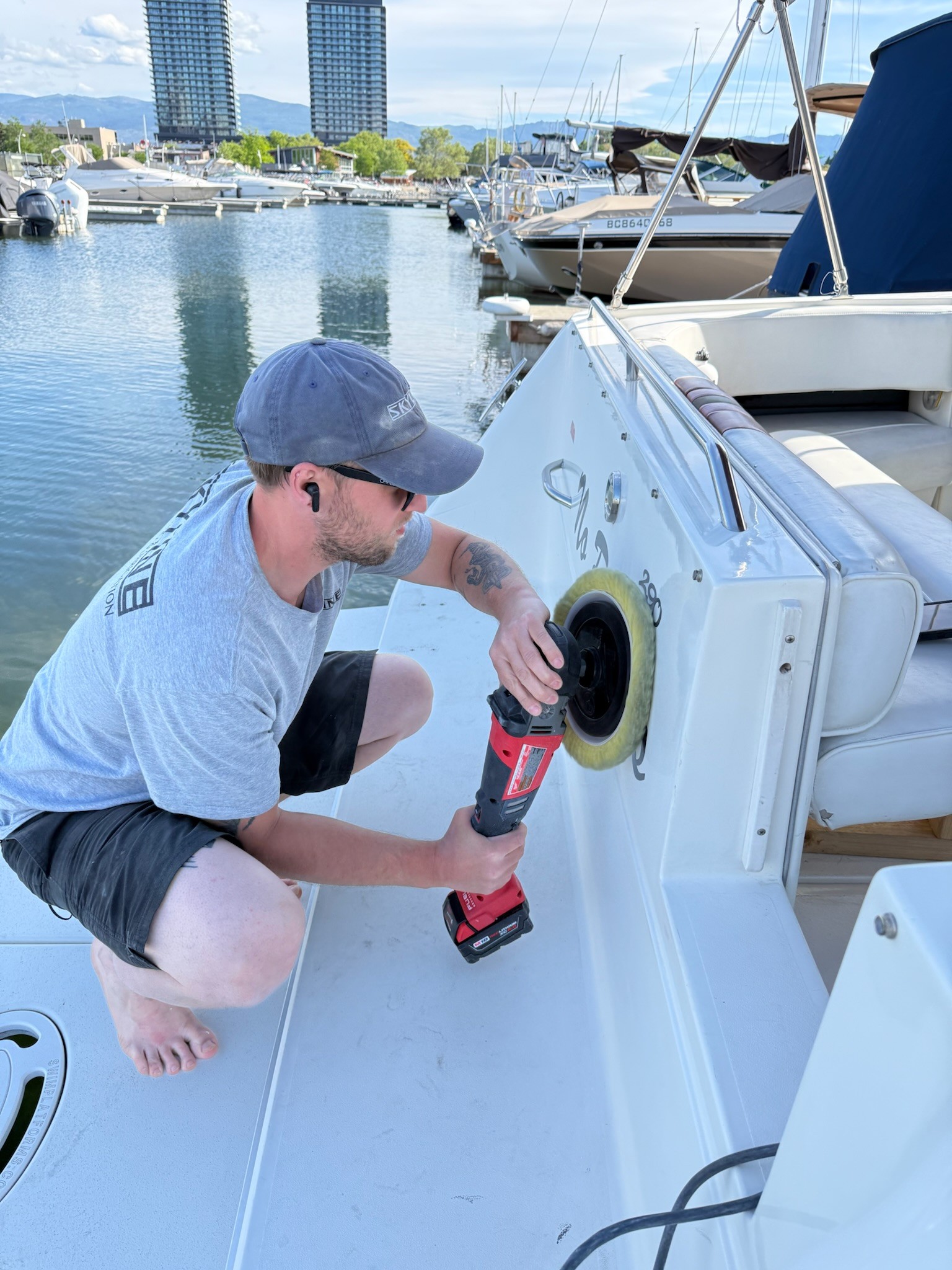 A person crouching down and working on getting into tough edges with their polisher. They are performing a heavy cut and buffing the marine detail to restore the boat