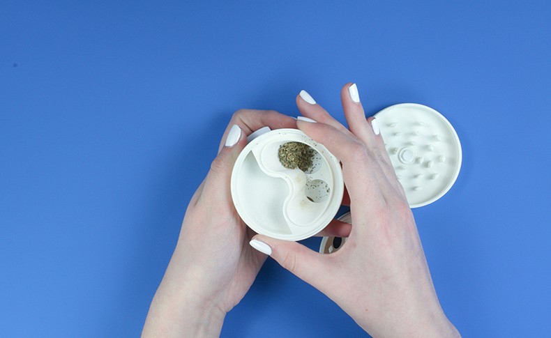 Two hands with white-painted nails are holding a white ceramic spice grinder against a solid blue background. The grinder has a lid with a textured surface and a separate compartment containing a dark brown spice. The hands are positioned as if preparing to use the grinder.