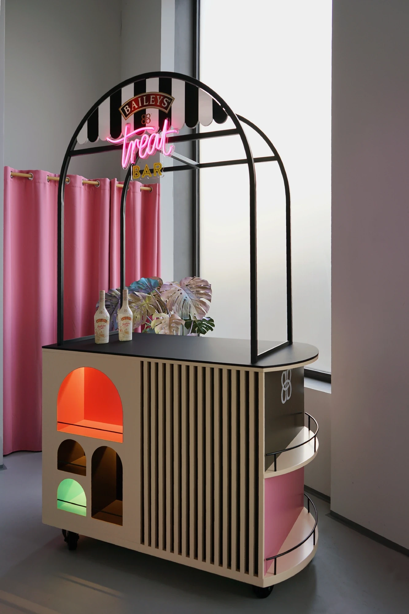 Baileys „Treat" mobile bar trolley with a black arched frame, pink neon signage, wooden body with carved arches and integrated coloured-light niches.