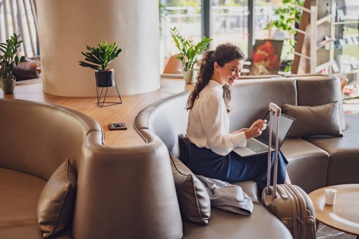 A woman sits in a cozy cafe, working on her laptop near large windows and comfortable seating.