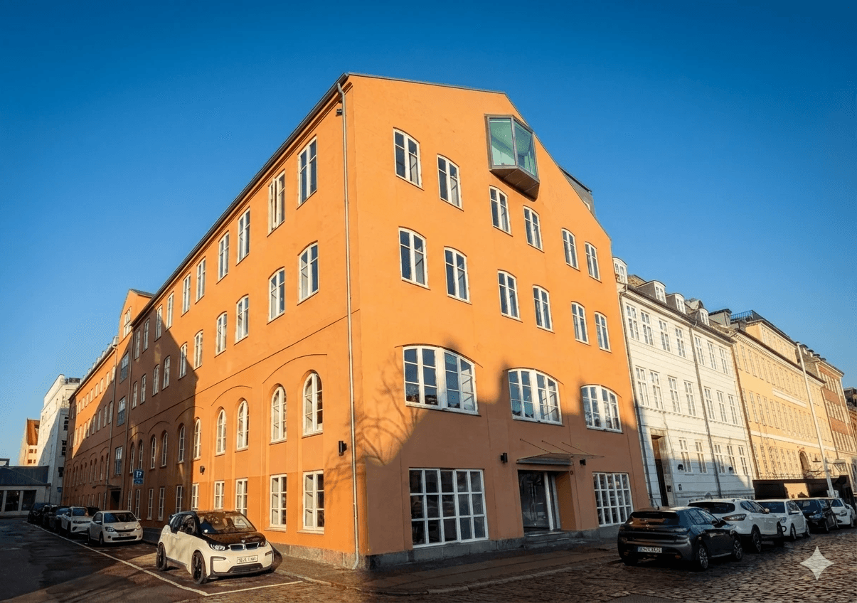 A green suspension bridge spanning across a body of water, with forested hills and residential homes in the background.