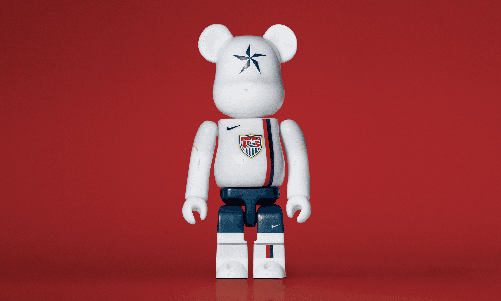A single 3.5” tall Bre@rbrick/Nike toy created to commemorate the 2006 US World Cup football team. The toy is painted to appear in the US football kit and stands in the center of the frame in front of a dark red background.