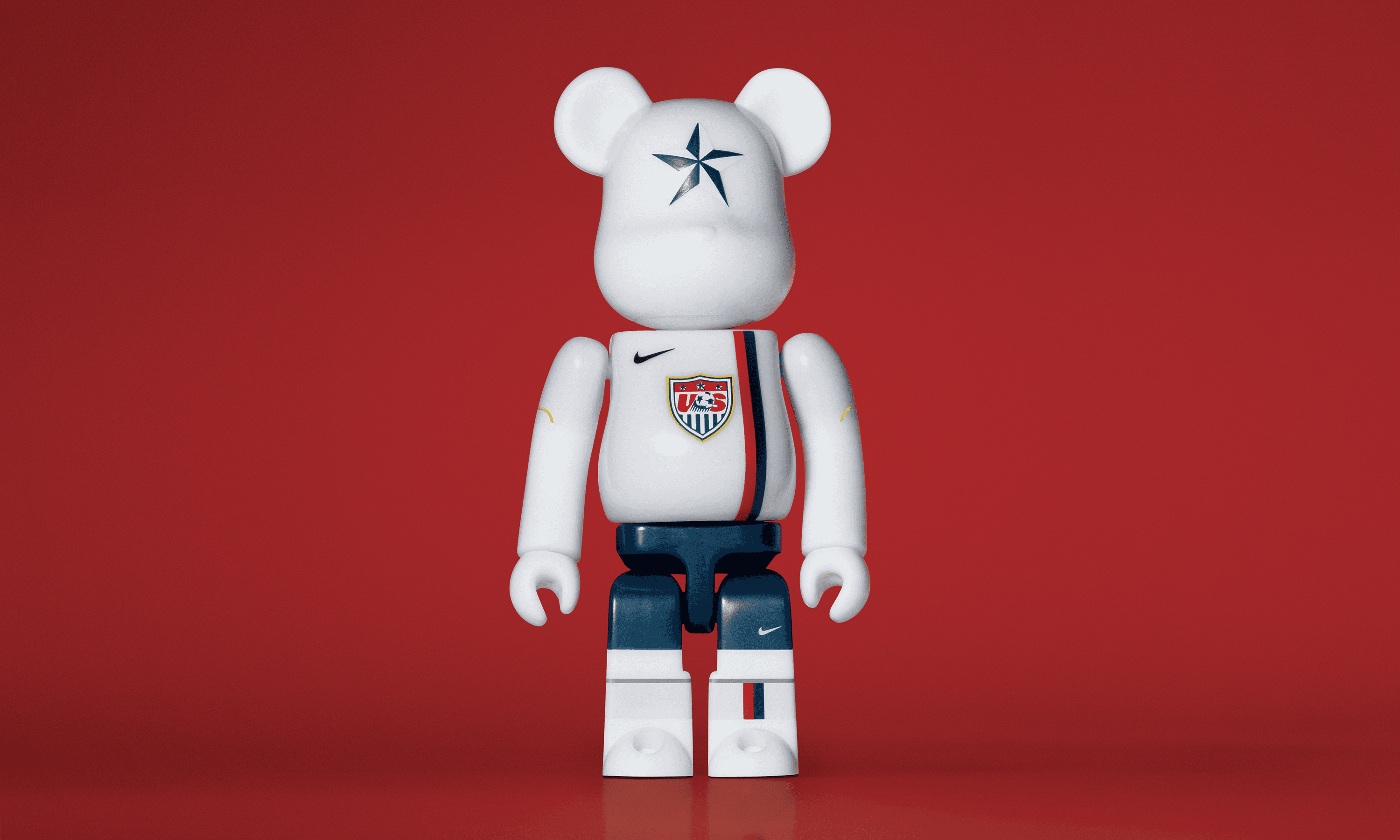 A single 3.5” tall Bre@rbrick/Nike toy created to commemorate the 2006 US World Cup football team. The toy is painted to appear in the US football kit and stands in the center of the frame in front of a dark red background. 