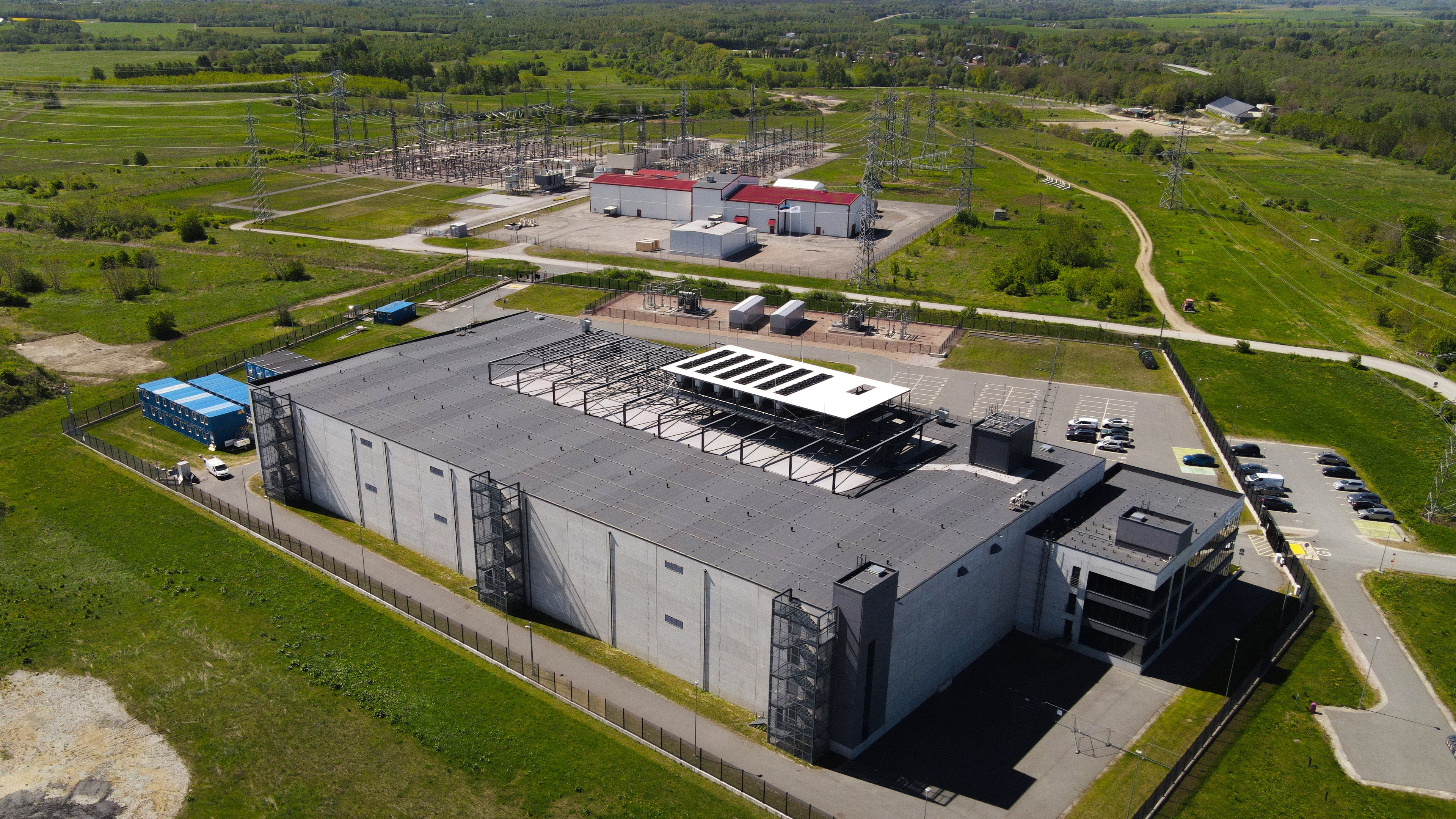Aerial view of a data center building on fenced land, with a substation behind it