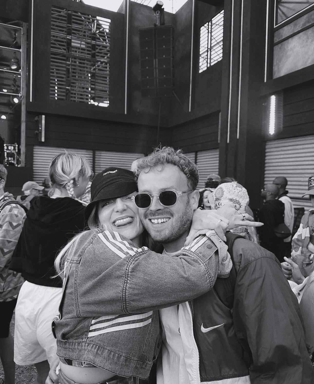 A couple stands on a sandy hill at sunset, facing away, with arms raised in a relaxed "hang loose" gesture. The scene conveys tranquility and joy.