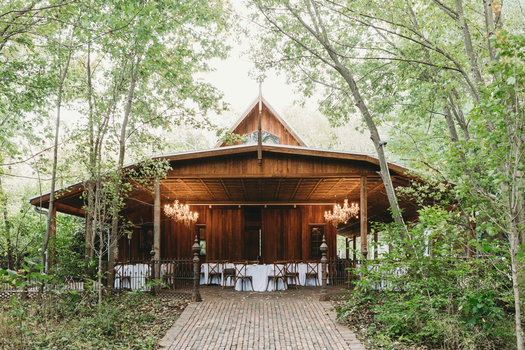 Rustic wooden wedding pavilion with crystal chandeliers nestled among tall trees along a brick pathway leading to the reception area