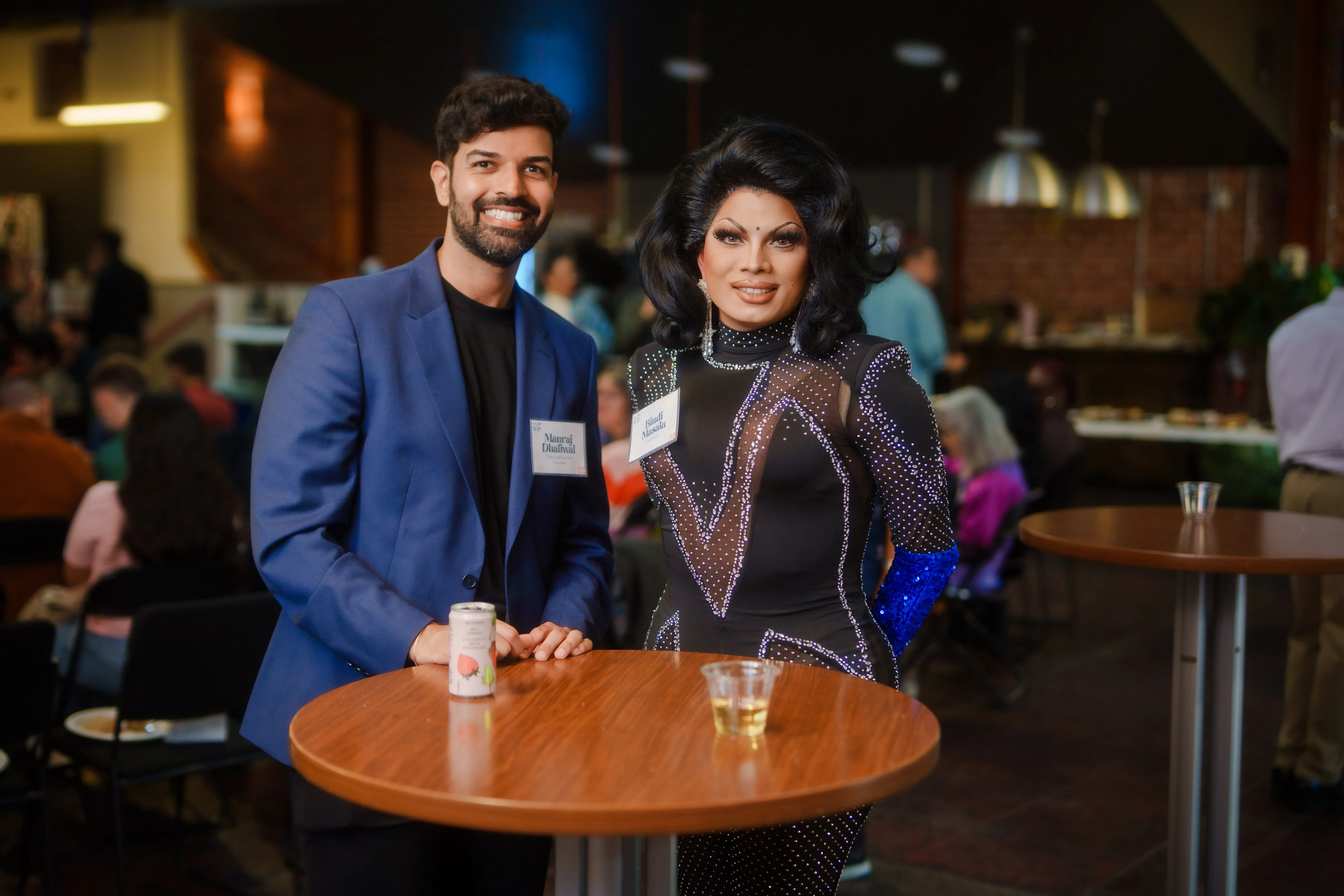 Uma pessoa de terno azul e outra com um vestido preto brilhante e maquiagem marcante estão sorrindo ao lado de uma mesa alta em um espaço de convivência social com pouca iluminação, com outros participantes e uma mesa de comida ao fundo.