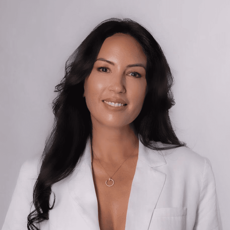 A young person with curly hair smiles at the camera while sitting in front of a laptop against a dark background.