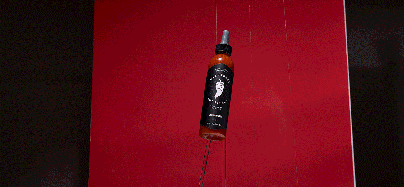 Hot sauce bottle suspended against a deep red backdrop, with visible compositing wires and dramatic lighting creating a floating product effect.