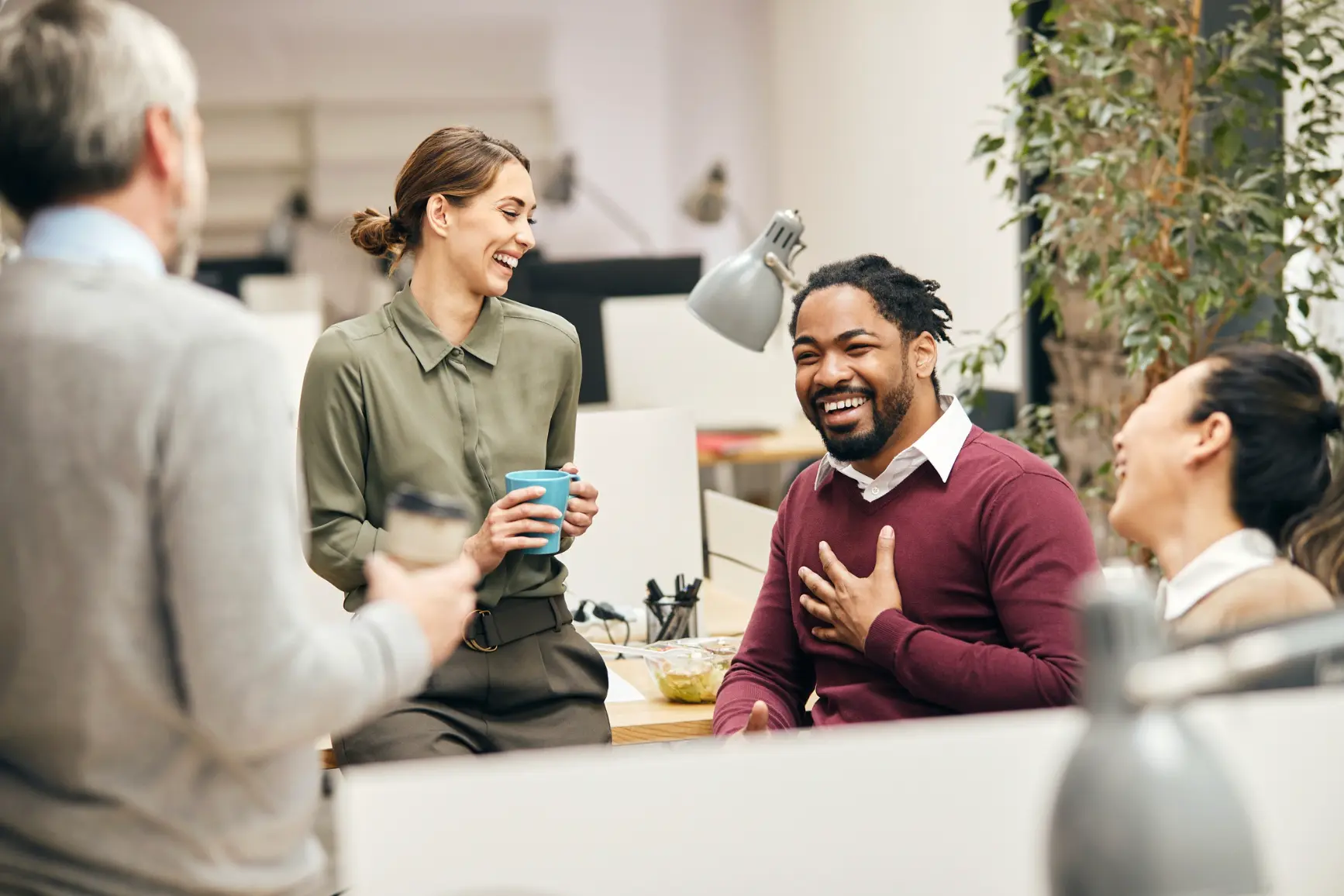 Diverse group of colleagues laughing together with coffee in a modern office, illustrating interpersonal connection and belonging at work