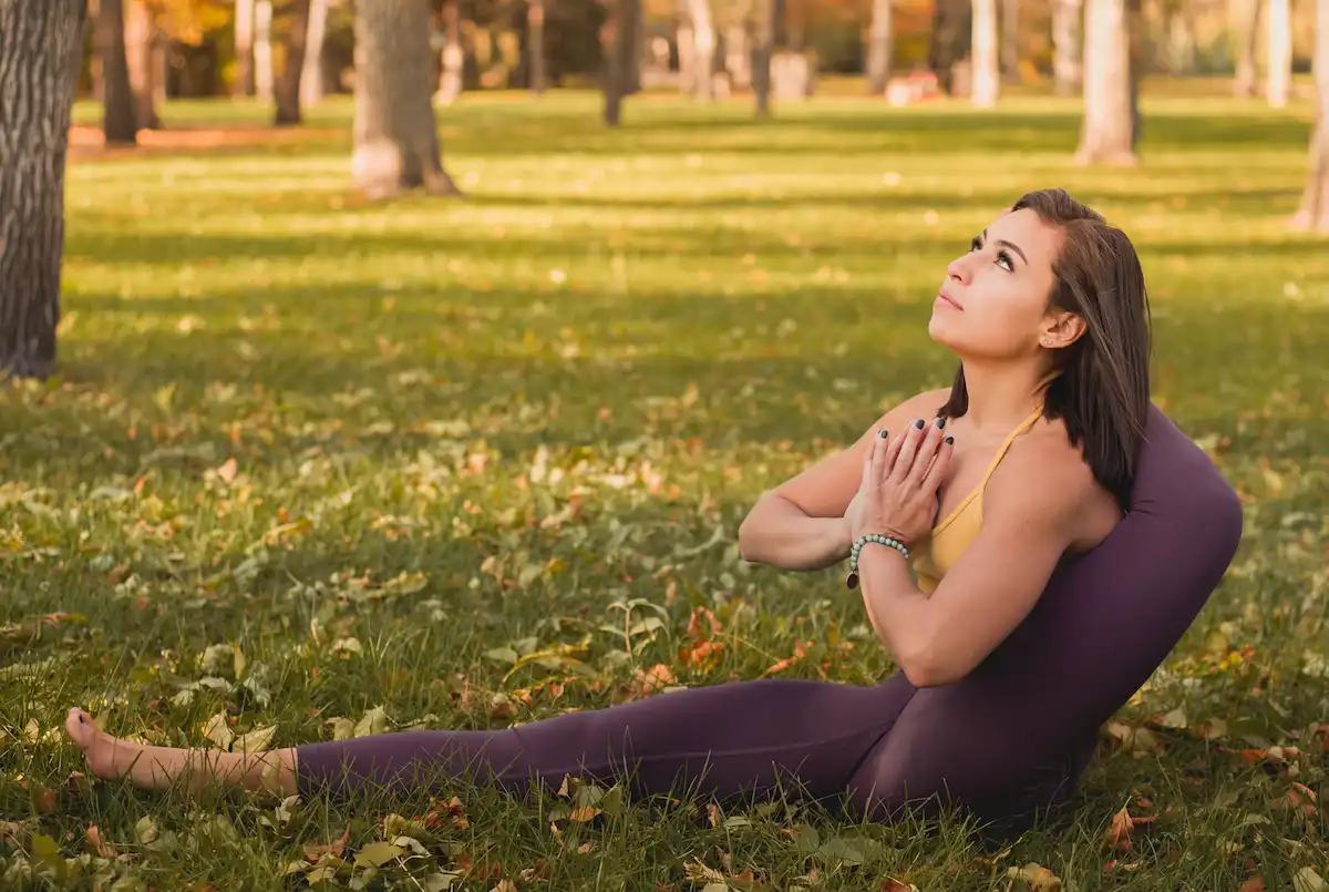 Woman practicing a seated spinal twist yoga pose with hands in prayer position on the grass outdoors.