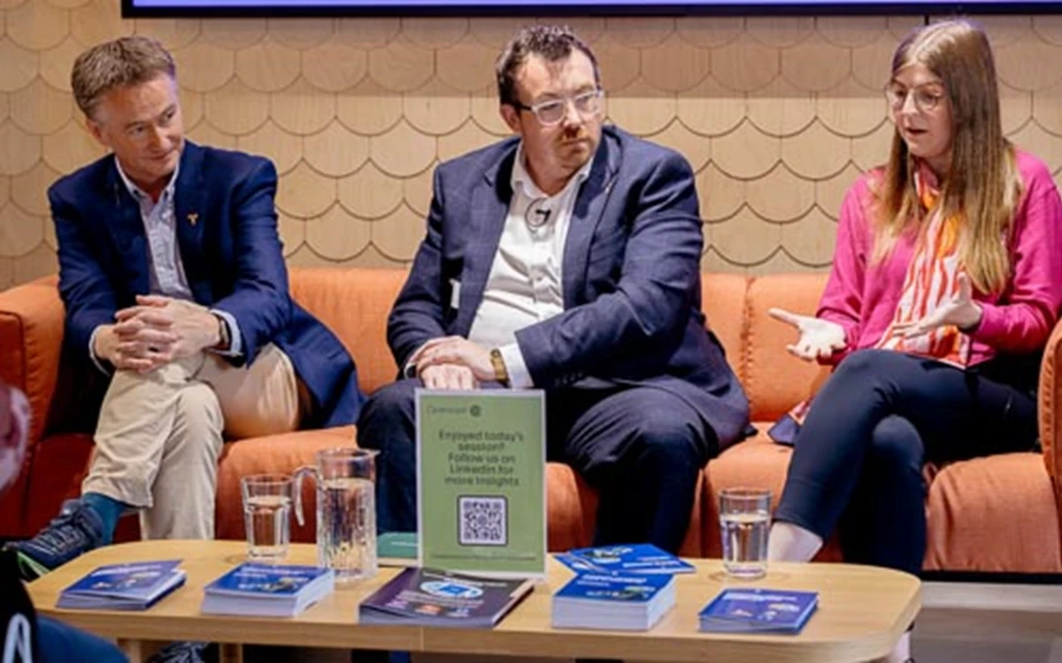 Three panel speakers sit on an orange sofa behind a low table with books, water glasses, and a sign displaying a QR code. The setting suggests a moderated discussion or talk at a public event.