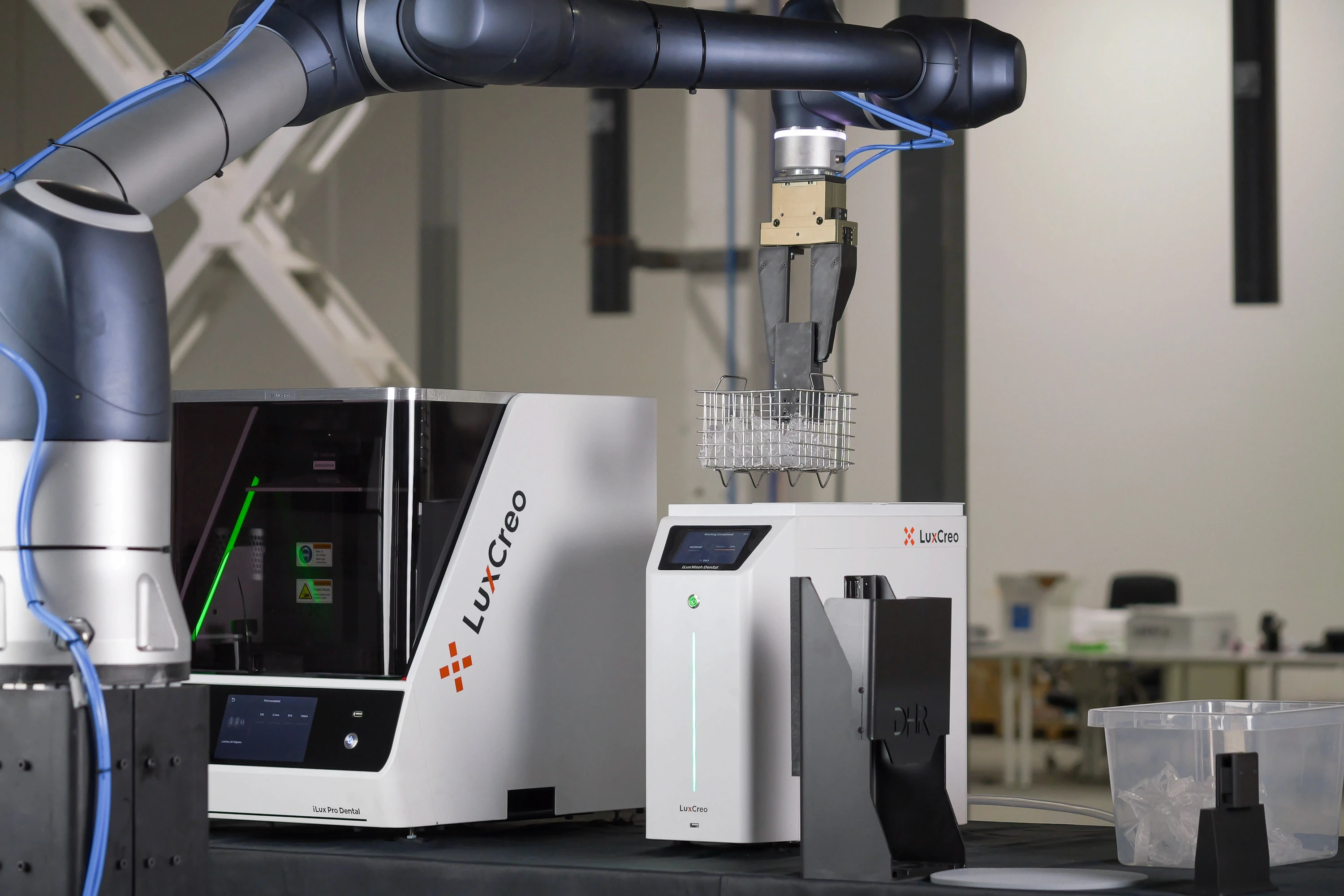 Robotic arm lowering a wire basket containing 3D printed dental parts into the iLuxWash Dental unit for automated cleaning.