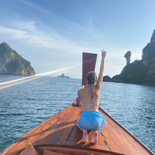 A person kneeling on a wooden boat, raising an arm towards clear blue waters and distant rocky islands under a sunny sky.