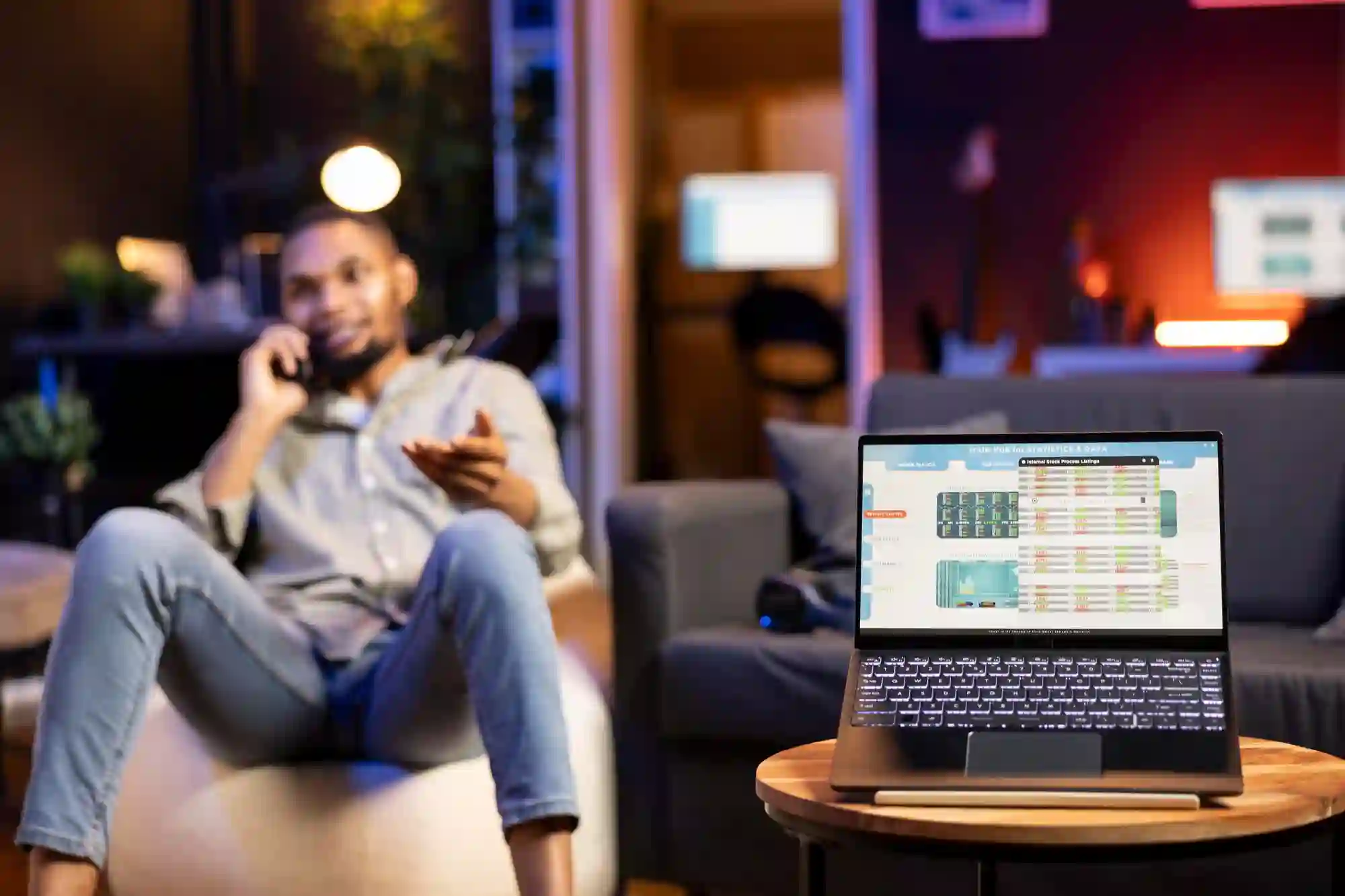Man talking on phone in a relaxed setting while a laptop displays business dashboard for remote work management.