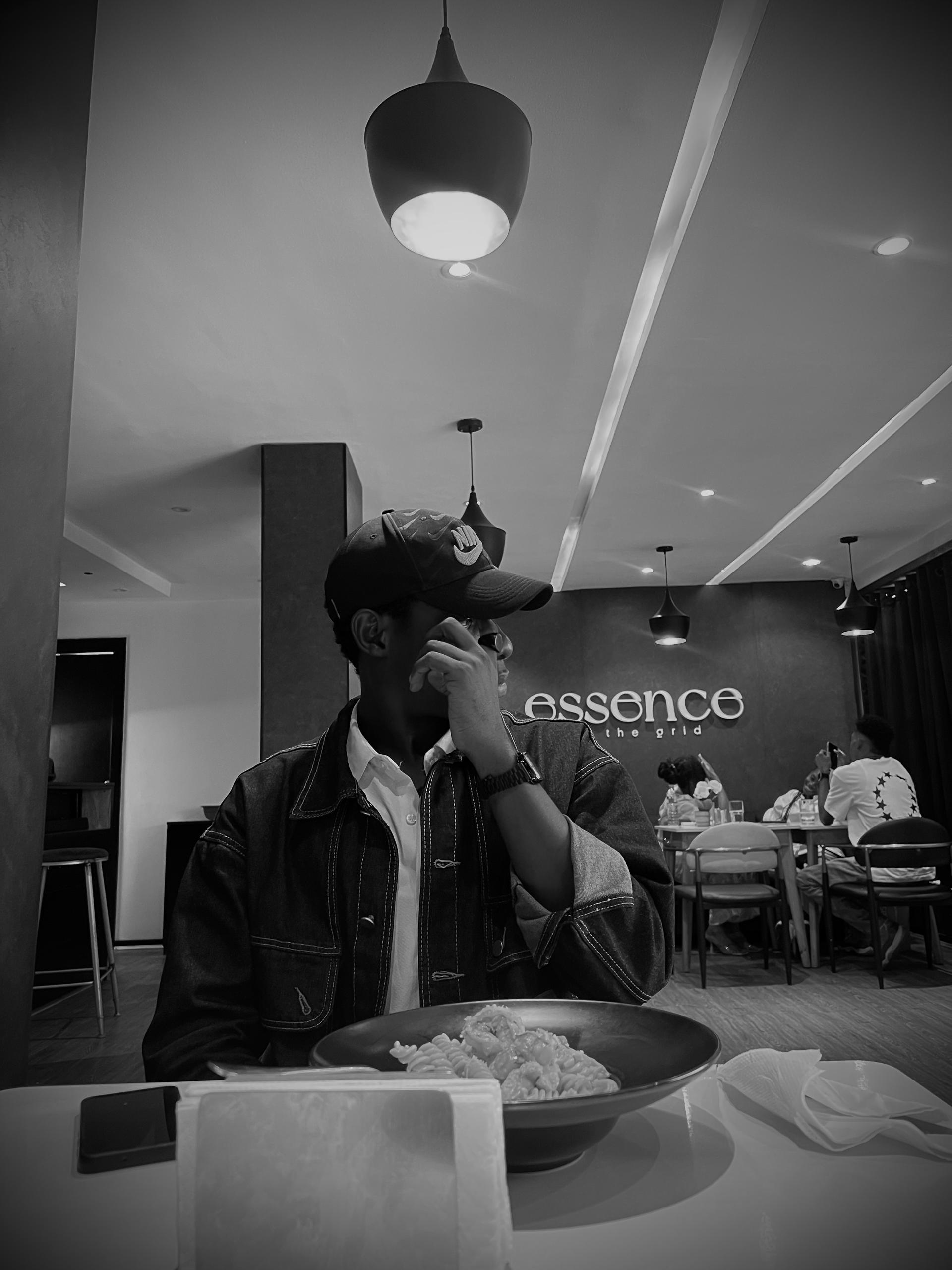 Person wearing black clothing carrying a black tote bag with a green globe-like geometric symbol, standing against a concrete wall in sunlight and shadow.