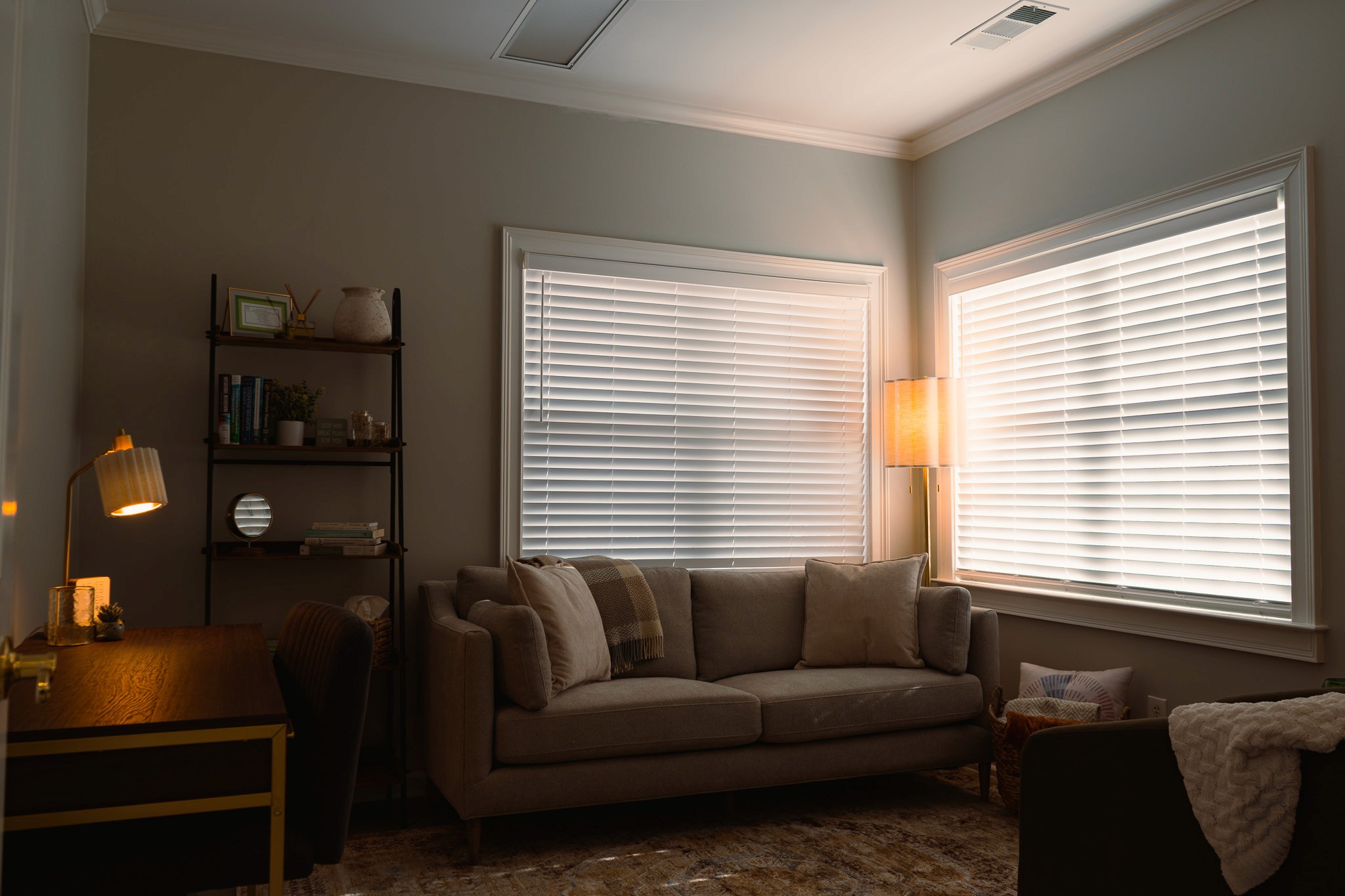 Therapy office with sofa, floor lamp, shelving unit, and window blinds with soft daylight.