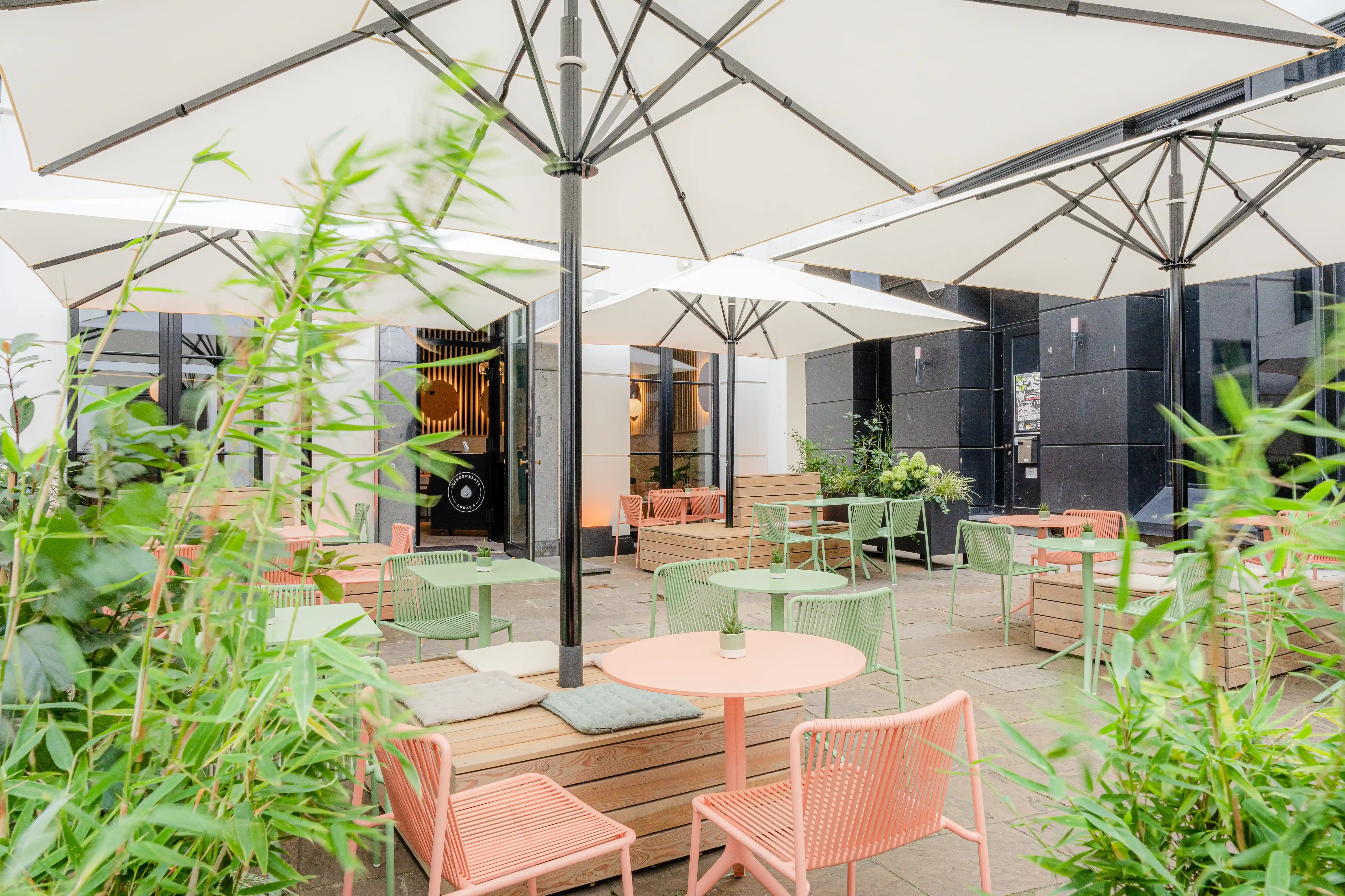 Terrace at LOKAL VIER with mint and coral wire furniture, round pink tables, wooden decks and benches under white parasols, framed by bamboo.