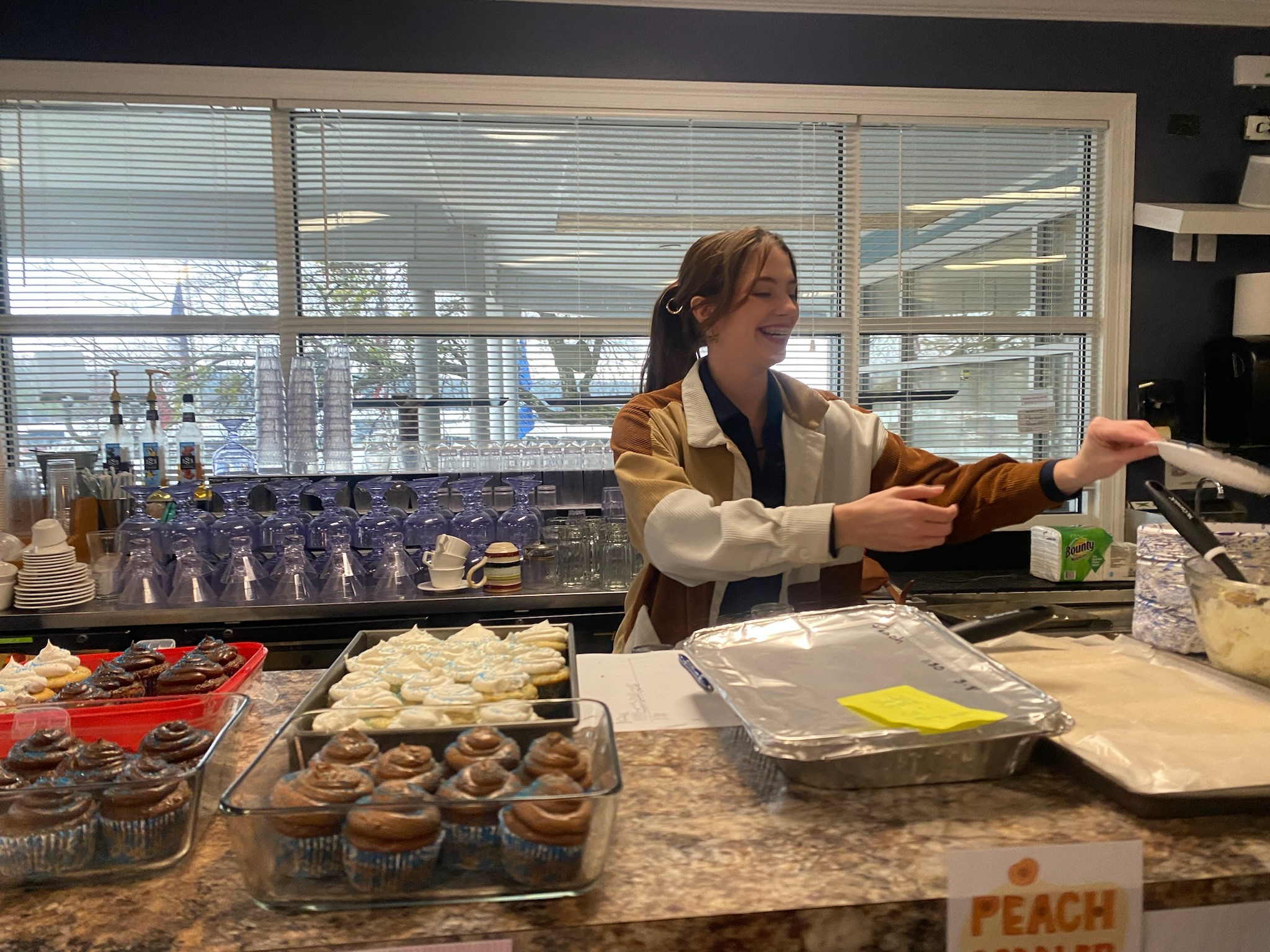 A cheerful barista with brown hair serves desserts and a hot drink behind a counter, which is adorned with trays of various cupcakes and pastries, against a backdrop of neatly stacked glasses and a large window letting in natural light.