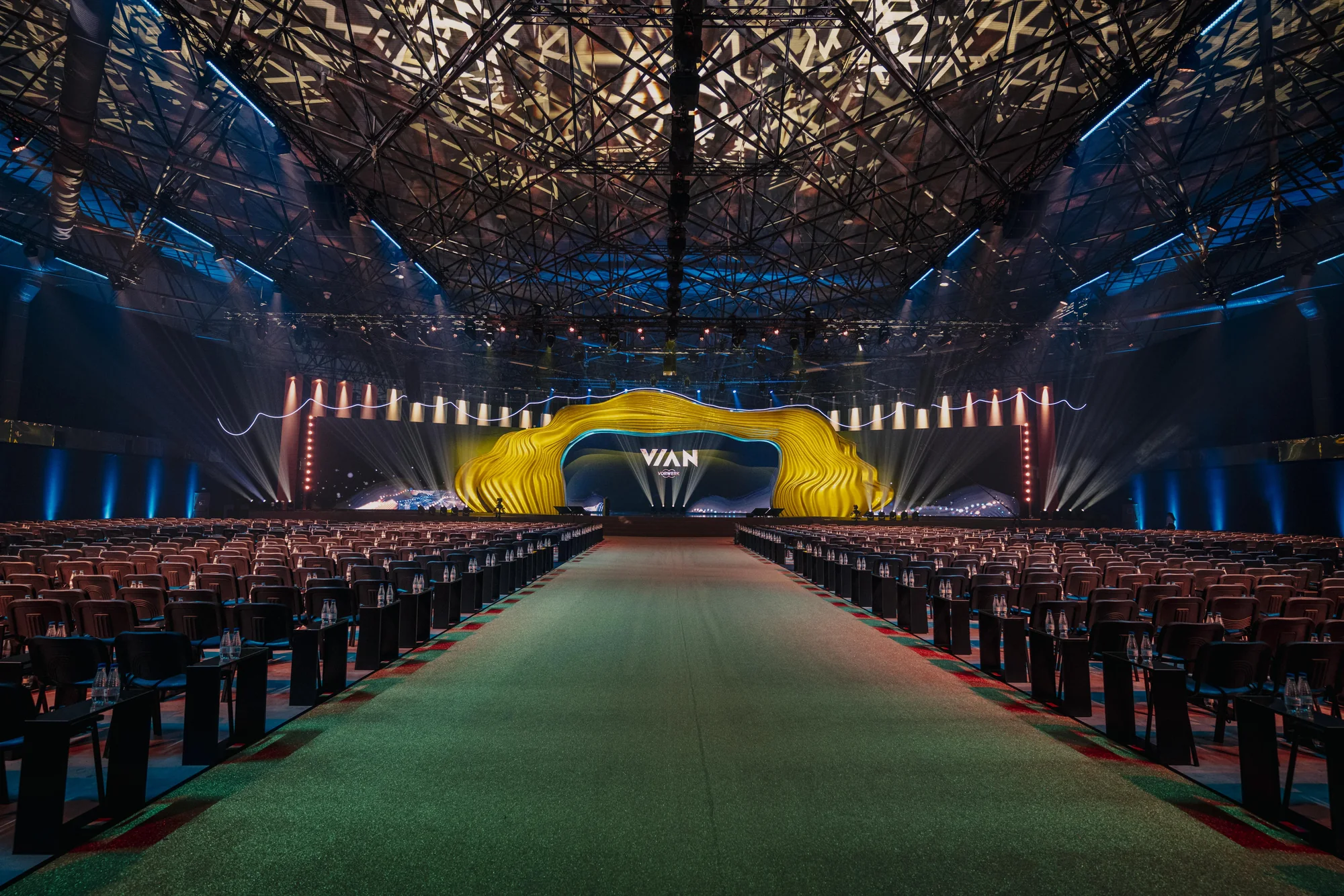 Vast event hall for Vorwerk VIAN 2025 featuring a sweeping yellow stage sculpture, rows of seating, and an illuminated green central aisle leading to the stage.