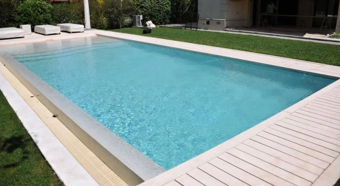 Moderna piscina rectangular de hormigón con terminación en Aquavations, bordeada por un deck de madera pintada de blanco y jardín verde.