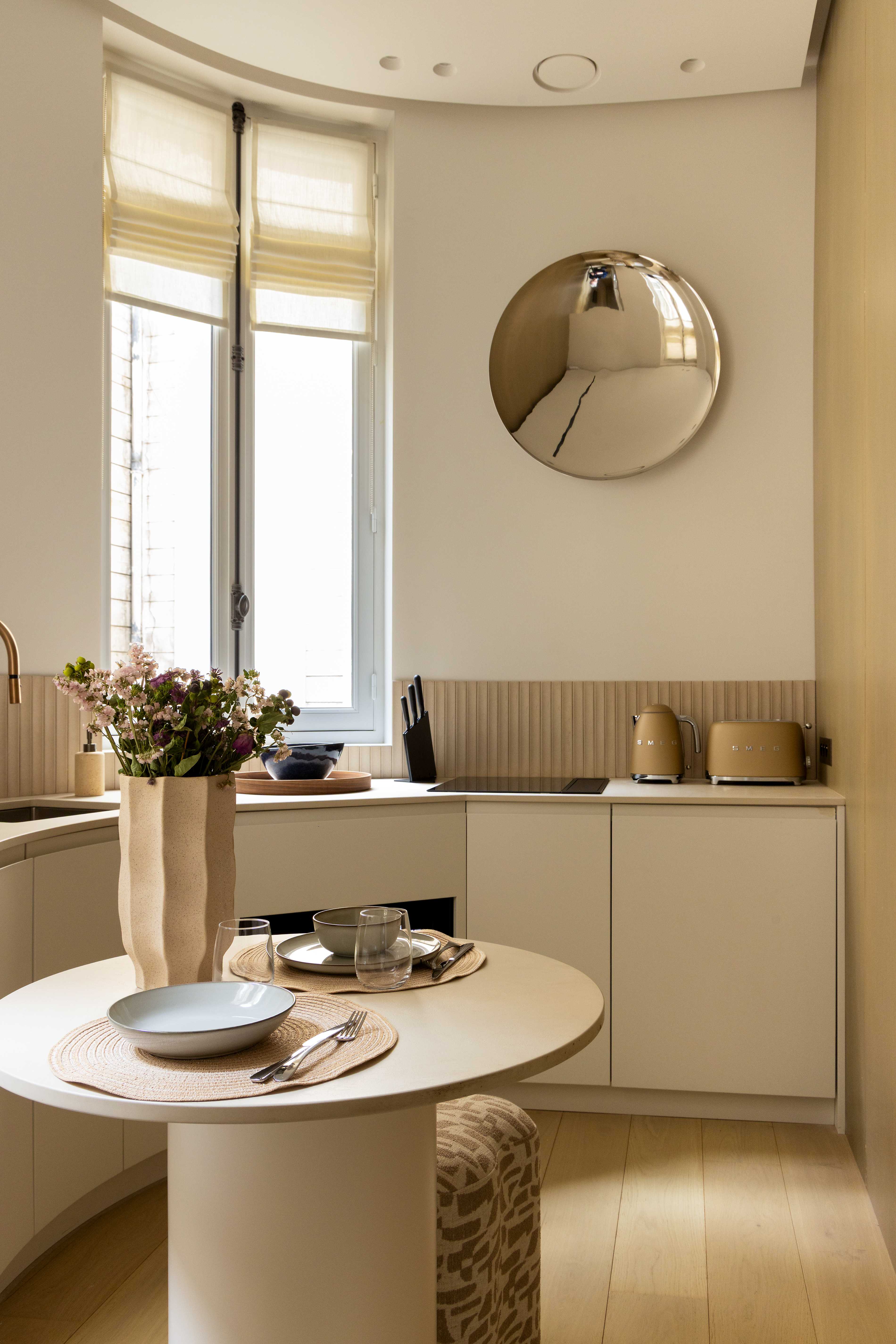 Kitchenette vue d'ensemble avec table ronde, miroir convexe chrome, credence cannelee et appareils Smeg dores