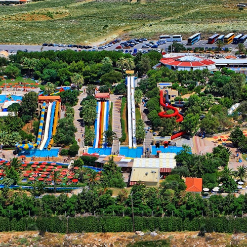 Aerial view of a water park featuring colorful waterslides, pools, surrounding greenery, and nearby buildings and parked vehicles.