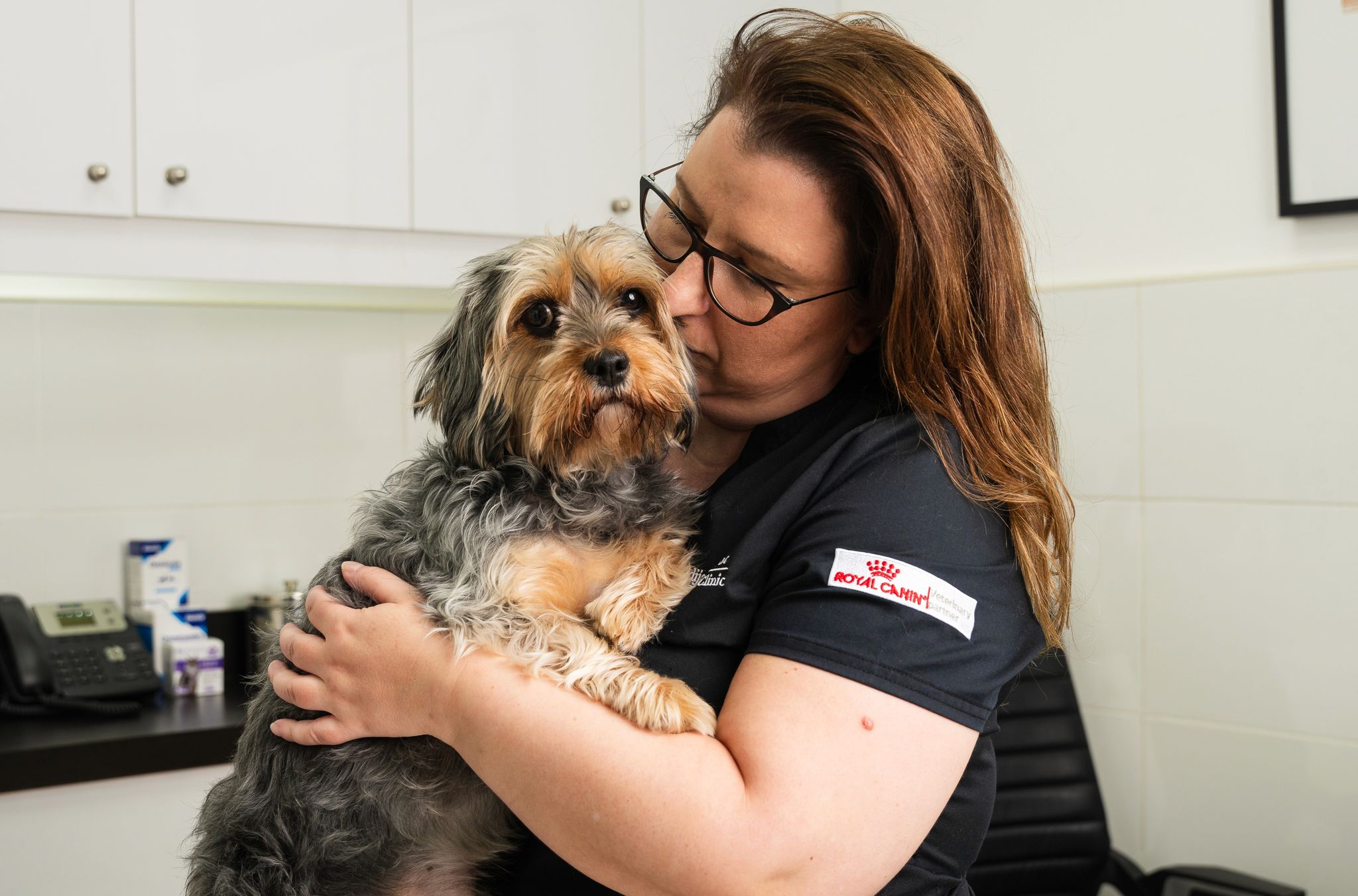 A female veterinarian in glasses lovingly hugs a small, fluffy dog in a veterinary office. The atmosphere is warm and caring, with pet supplies in the background.