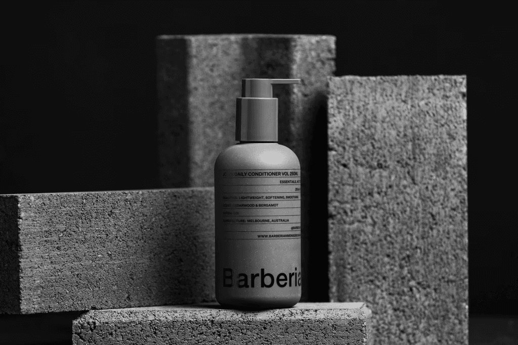 Black-and-white product shot of a Barberian Men's Grooming bottle, displayed against a backdrop of textured concrete blocks.