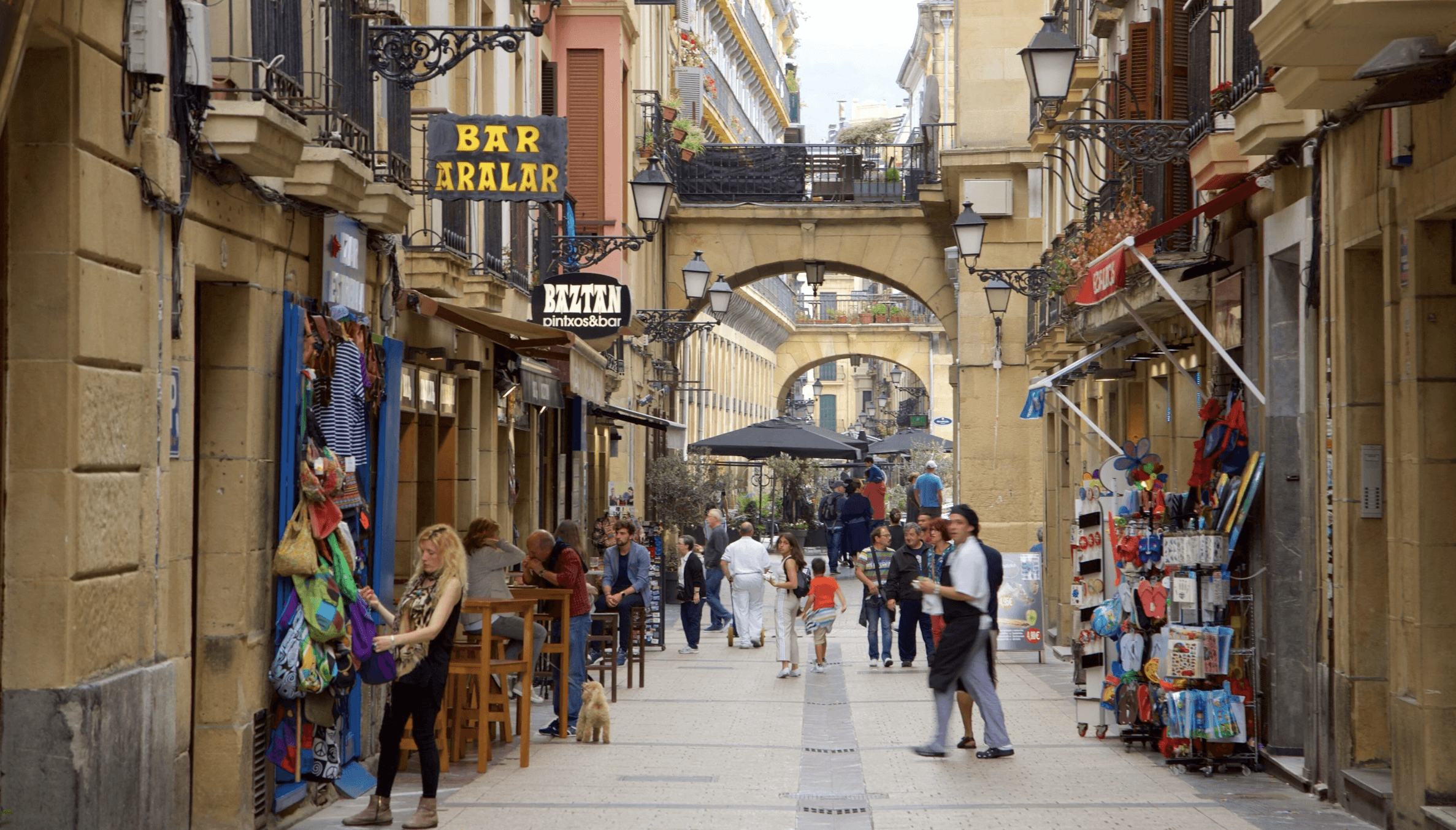 A person walks along a seaside pathway, gesturing with one hand, wearing a jacket and casual shoes.