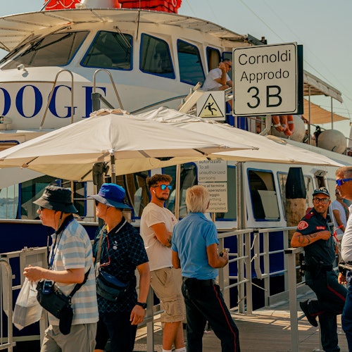 在白色遮阳伞下，人们走在 Cornoldi Approdo 3B 一艘停靠的蓝白相间船只附近。
