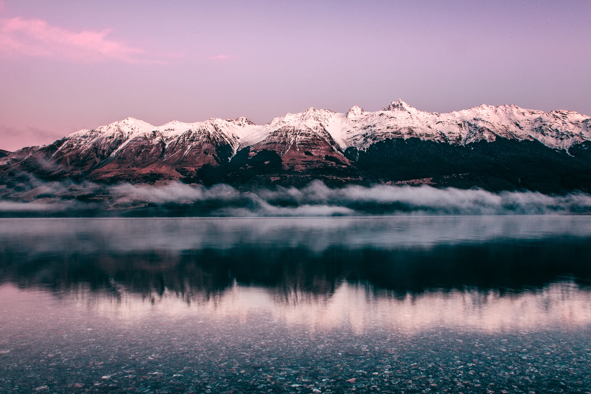 Snow-capped peaks above low mist and a still lake under a pink dawn sky