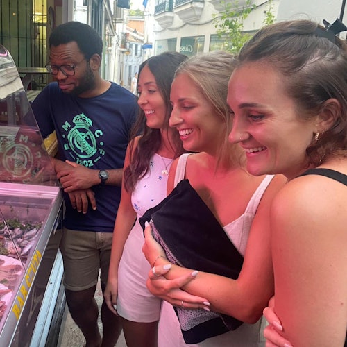 Four people smiling and looking at food displayed in a street market counter on a narrow, brightly lit street.