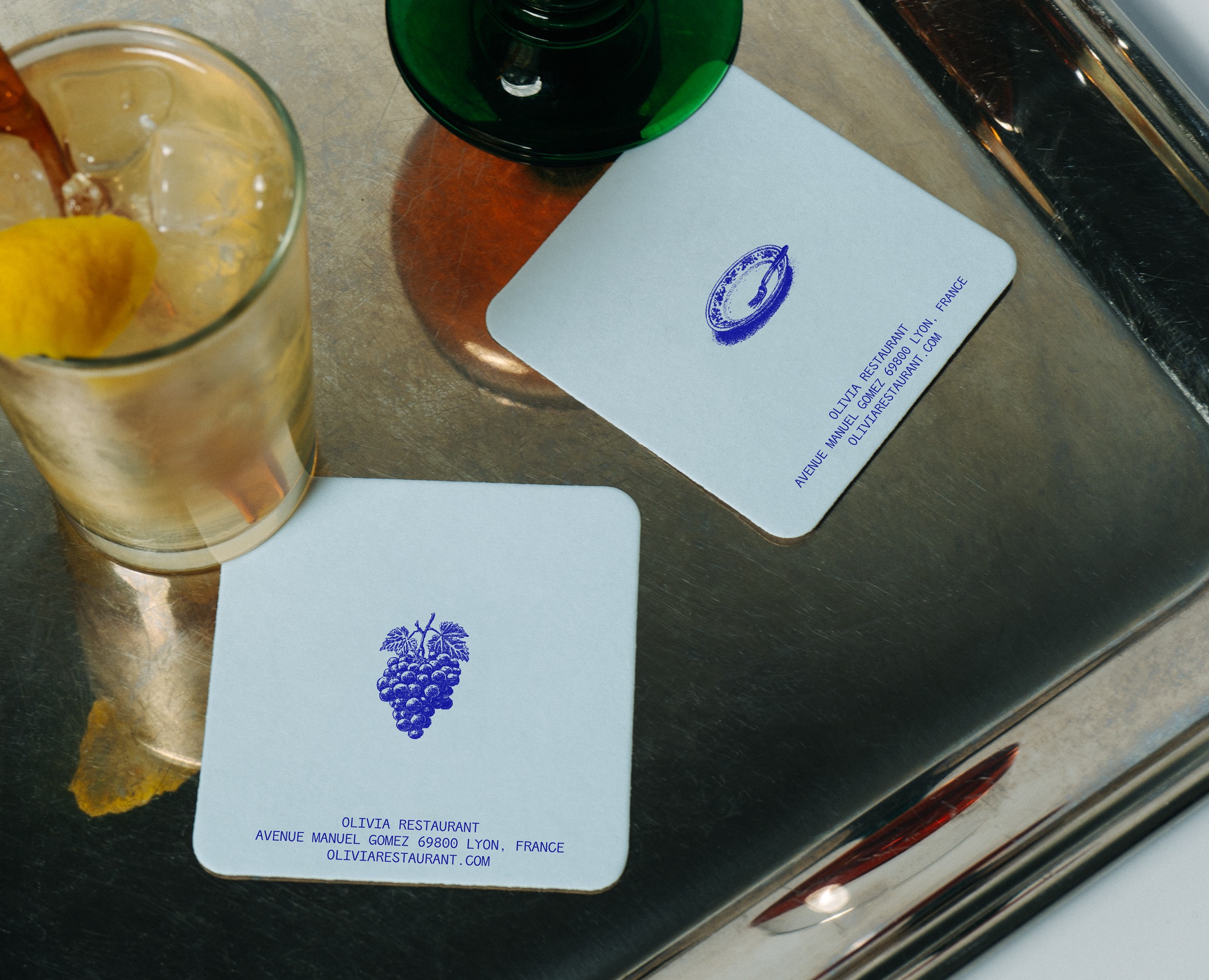 Sous-verres carrés OLI•VIA posés sur un plateau en métal avec cocktails, illustration bleue de poulpe pour restaurant de fruits de mer méditerranéen.