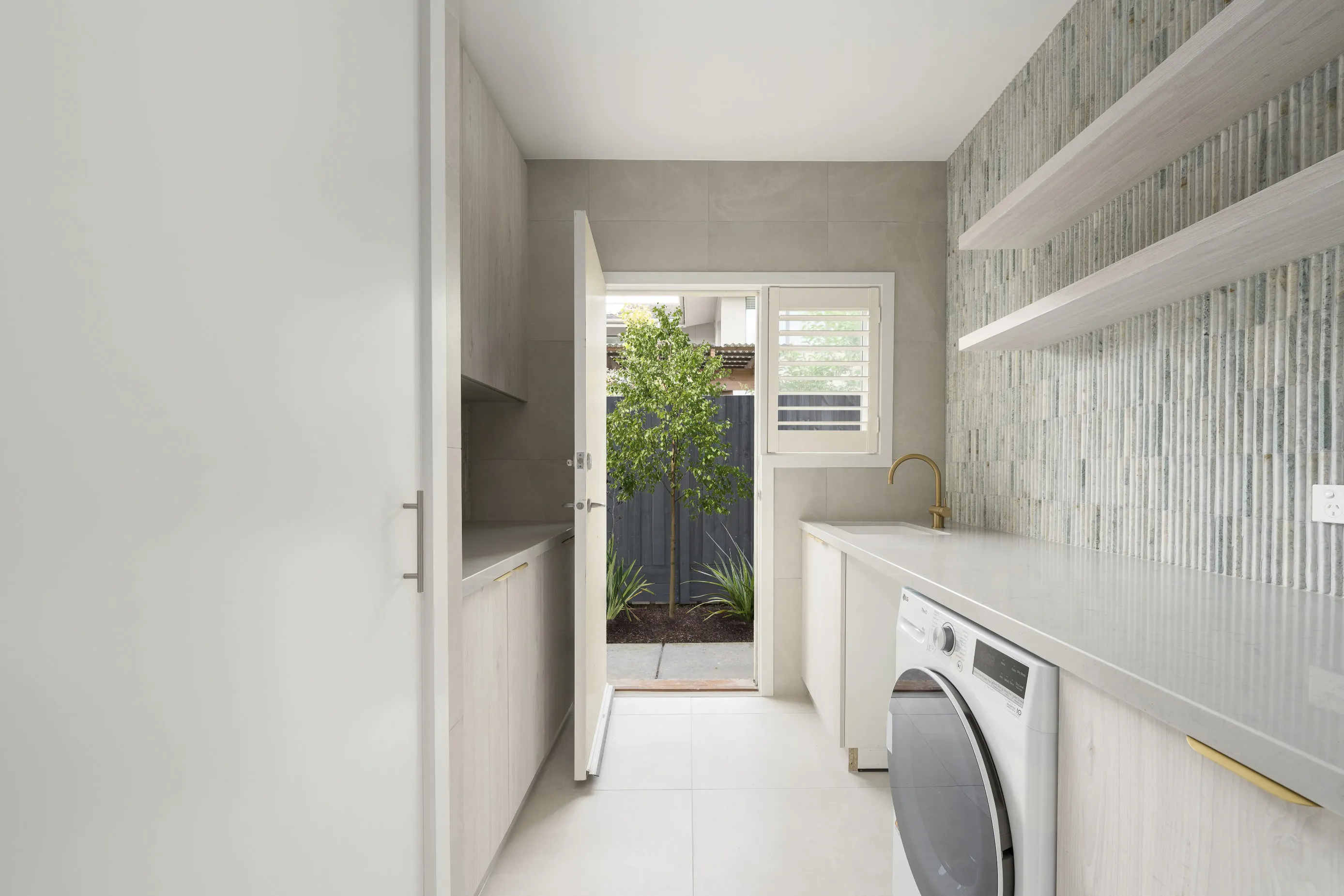 Bundoora laundry with whitewashed oak cabinetry, stone benchtop, fluted tile splashback and a door opening to a planted courtyard