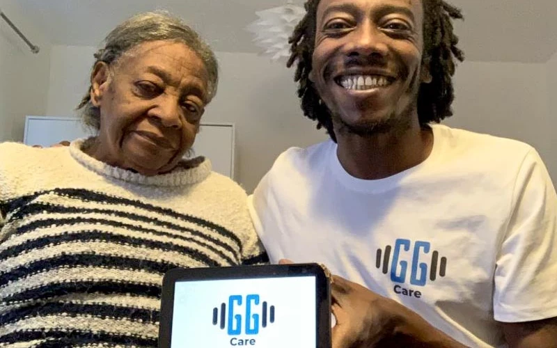 Two people sit indoors, holding a tablet that displays the “GG Care” logo. One person wears a striped sweater, and the other wears a white T‑shirt with the same logo printed on it. The setting appears casual, with a plain interior background.