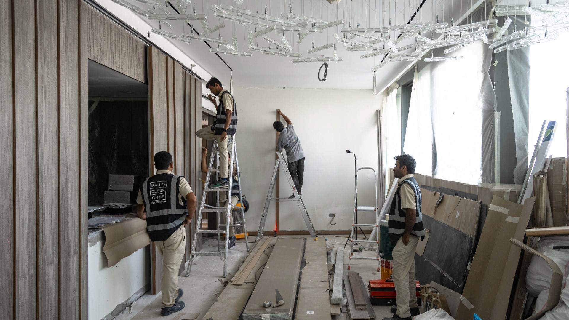 Four workers in uniform install ceiling tiles and paint walls in a partially constructed room, surrounded by tools and building materials.
