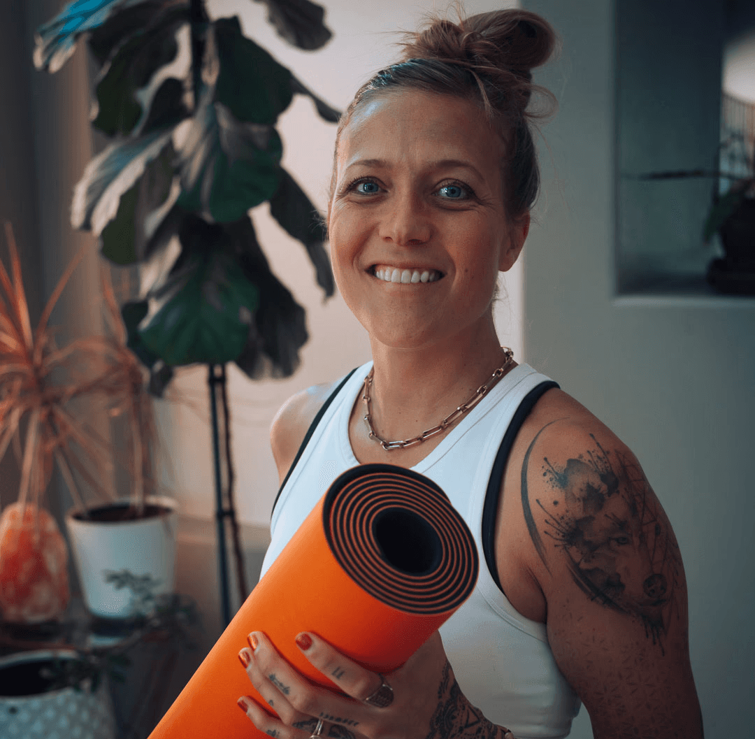Bre holding an orange yoga mat in a room with plants. She wears a white tank top, and exudes a calm, positive vibe.