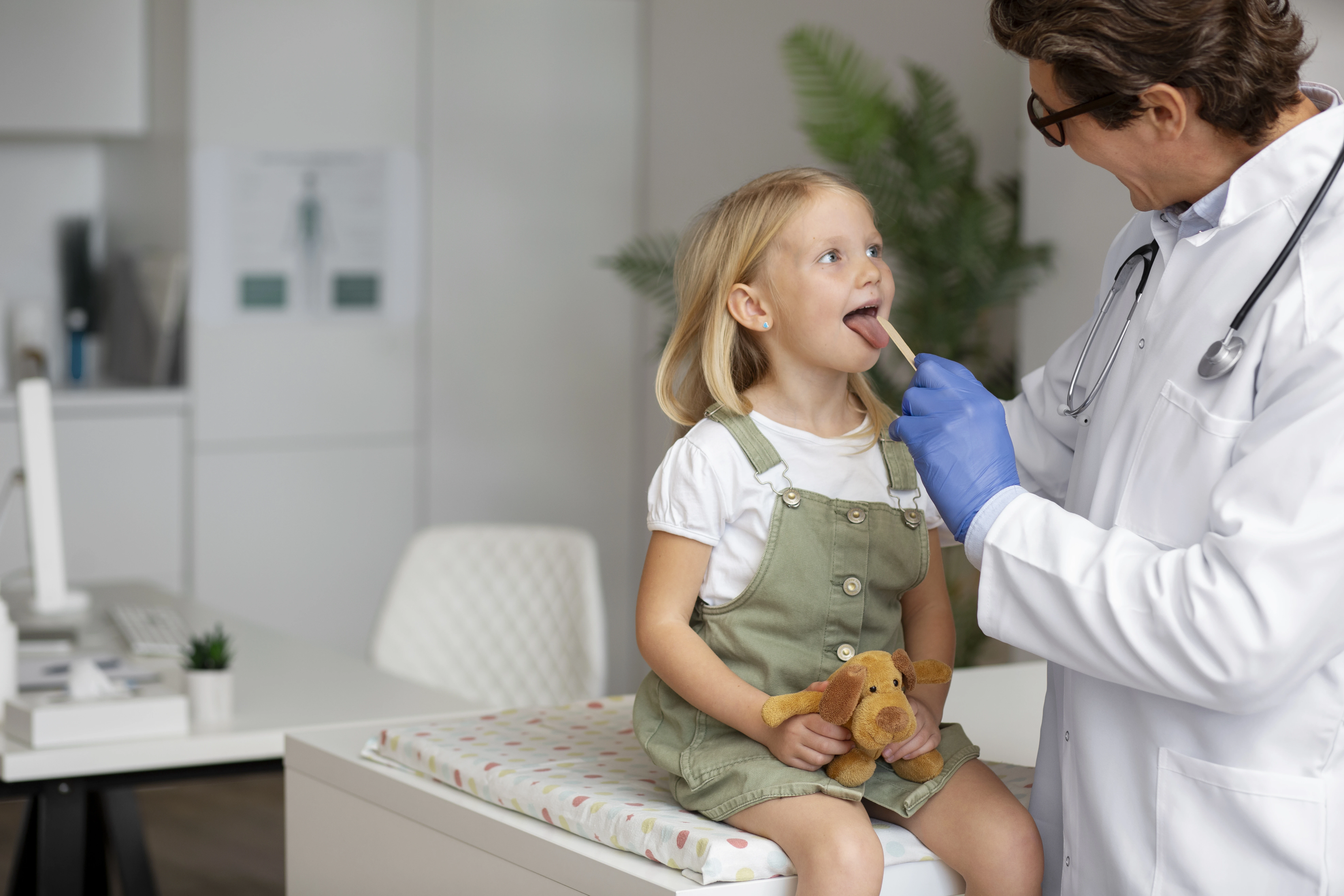 Um médico (pediatra) com óculos e estetoscópio, usando luvas azuis, examinando a garganta de uma menina loira sentada em uma mesa de exame. A menina segura um ursinho de pelúcia marrom.