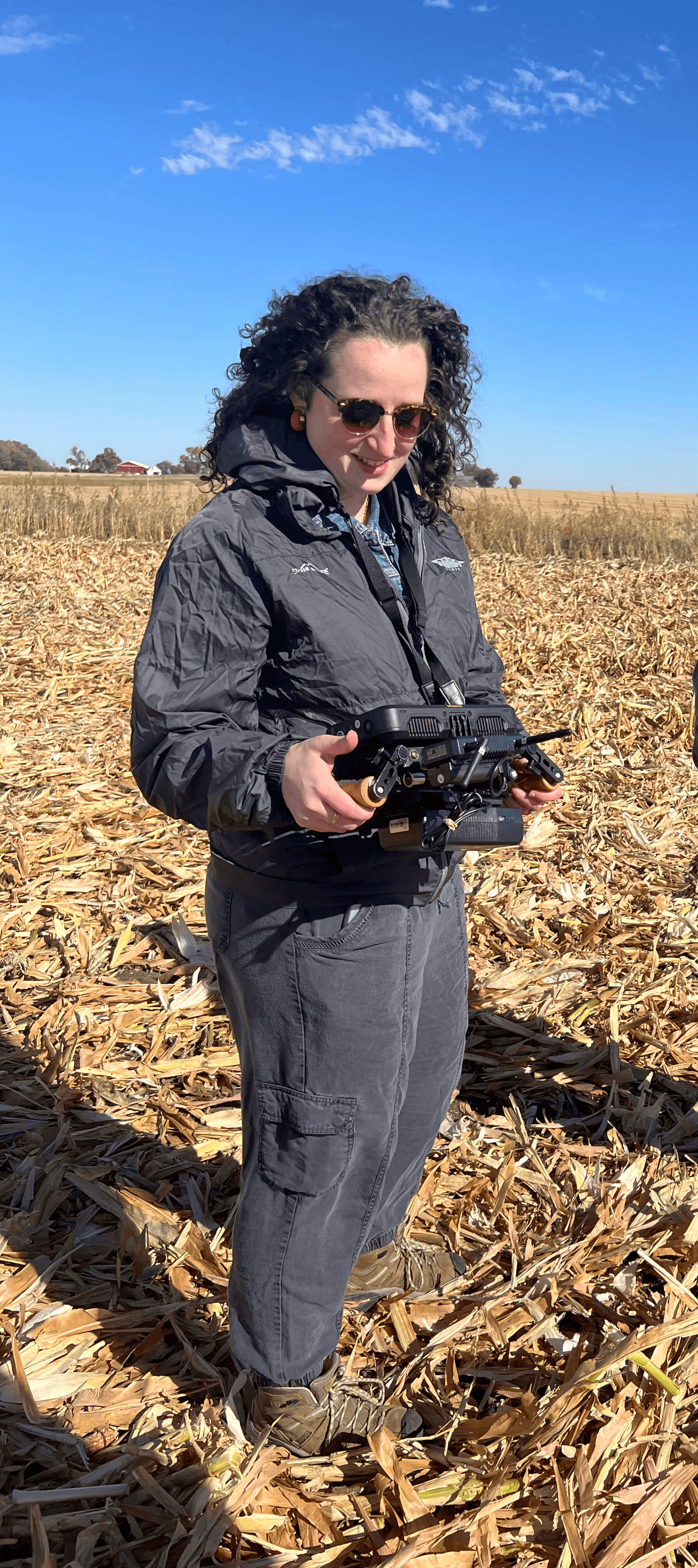 A woman stands in a just-plowed cornfield, looking at a remote viewfinder that shows her the footage a drone is capturing.