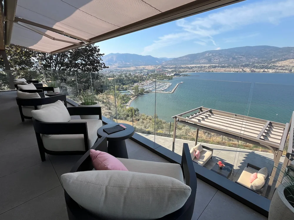 Luxury patio with frameless glass railing overlooking expansive lake and mountain views, featuring lounge seating and overhead awnings.