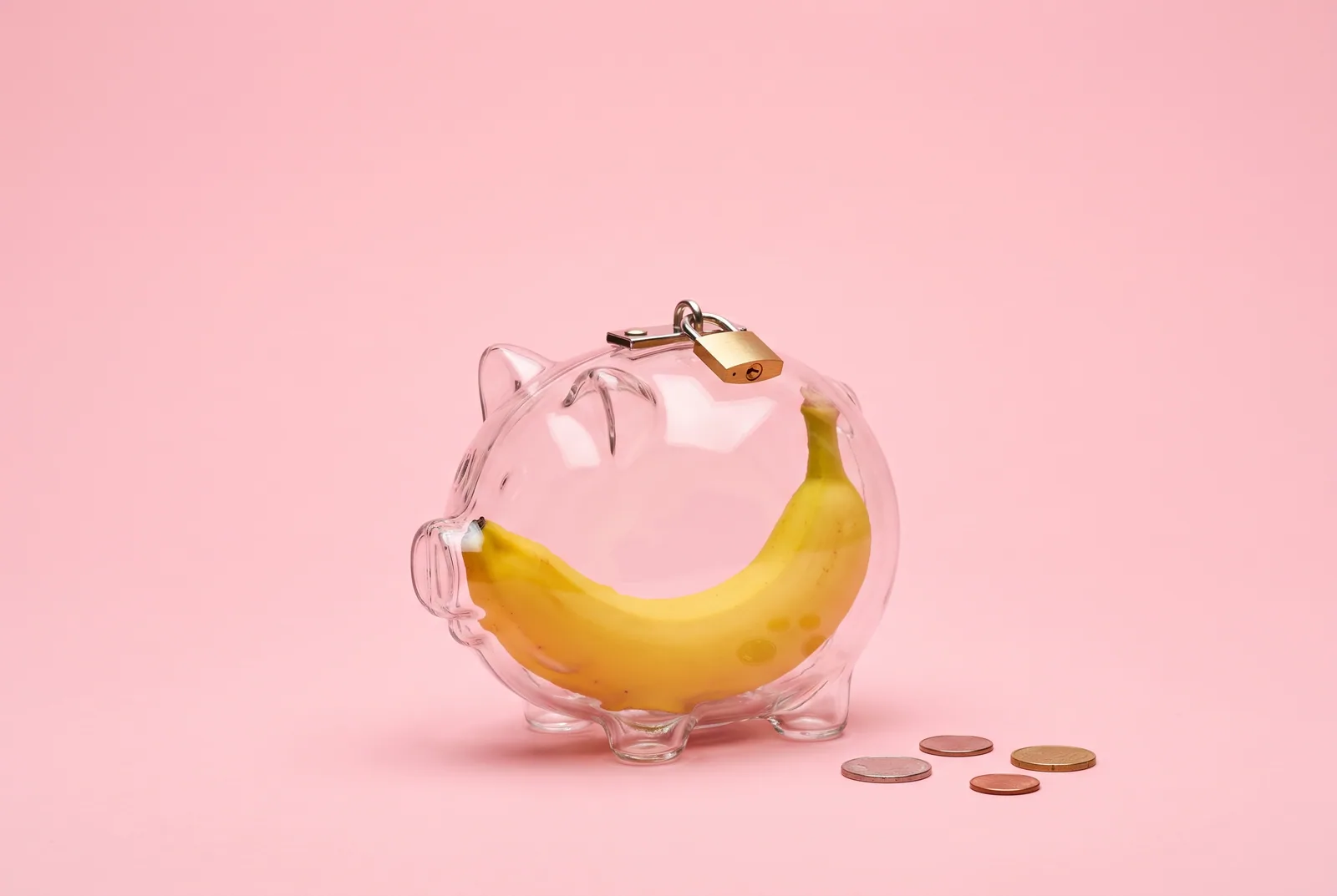 How to stop spending money on OnlyFans concept: a banana locked inside a transparent piggy bank with a small brass padlock on a pastel pink backdrop