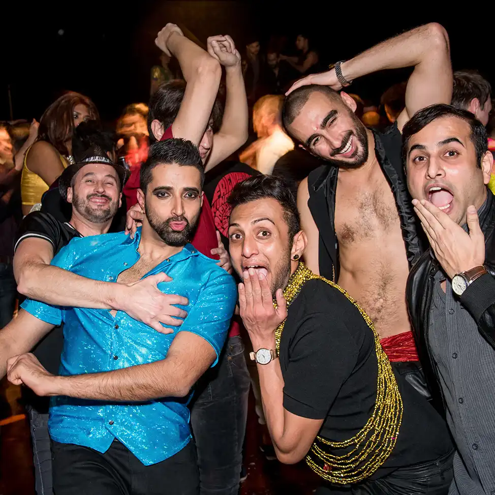 A lively group of five men pose playfully at a crowded party. They strike funny faces with arms around each other, exuding joy and camaraderie.