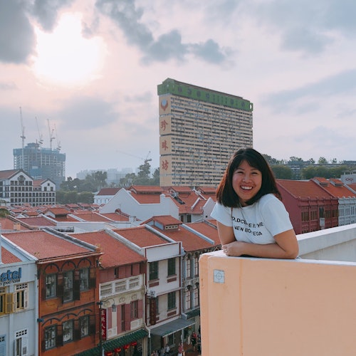 一位女士在屋顶微笑，背景是一片红瓦建筑和一幢带有中文标识的高楼，天空中布满云层。