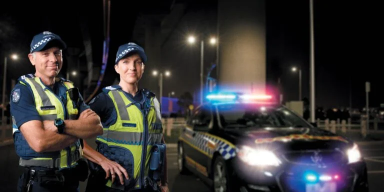 Two Victoria Police officers in hi-vis vests beside a police car with flashing lights at night, 'Be a force for good'