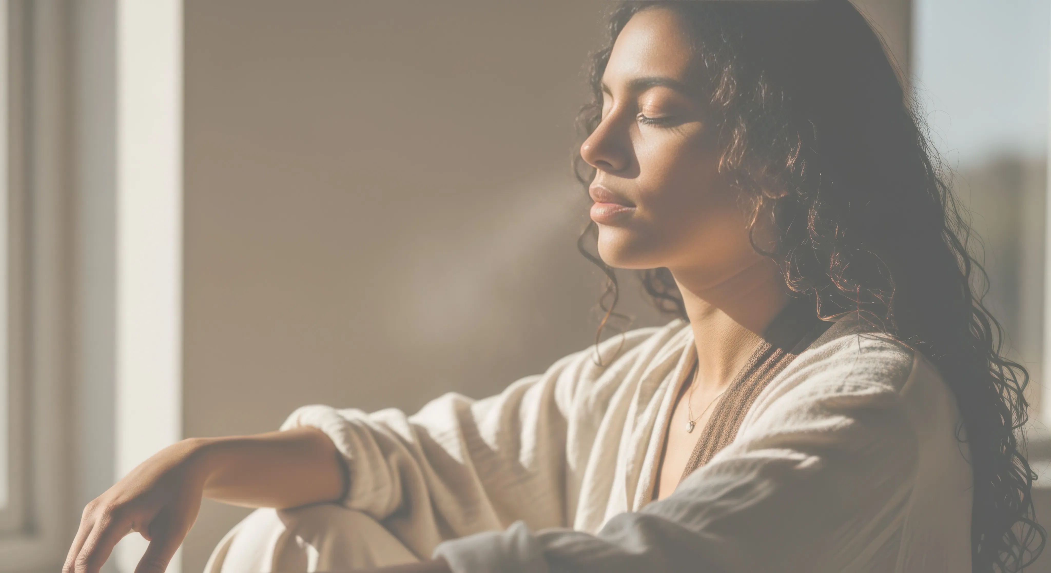 A woman with curly hair sits indoors in soft sunlight, eyes closed, wearing light-colored clothing and a necklace, appearing calm and relaxed.