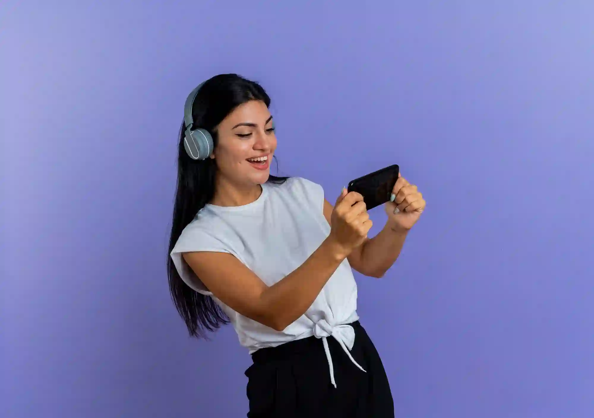 A smiling young woman wearing headphones holds a smartphone horizontally, looking engaged, against a purple background.